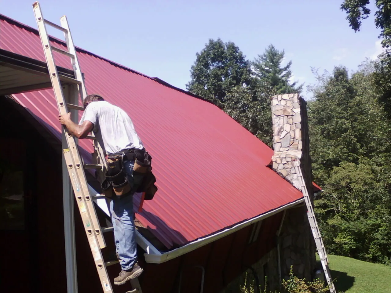 Roofer on a ladder beside a red metal roof and stone chimney — Soffit & Fascia Repair by Roofing Brothers in Galesburg
